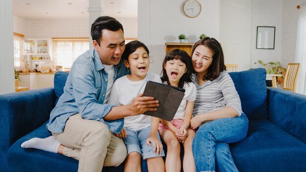 Papá de familia asiática alegre feliz, mamá y niños divirtiéndose y usando videollamadas de tableta digital en el sofá de la casa.