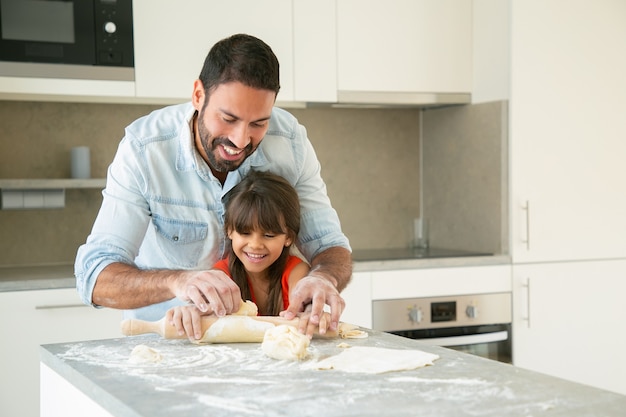 Papá feliz y alegre y su niña disfrutando del tiempo juntos mientras amasan y amasan la masa en la cocina.