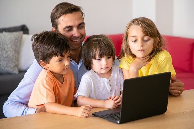 Papá feliz y niños viendo películas a través de la computadora portátil juntos. Padre caucásico sentado a la mesa y abrazando a niños lindos. Niños y niñas mirando la pantalla. Concepto de paternidad y tecnología digital