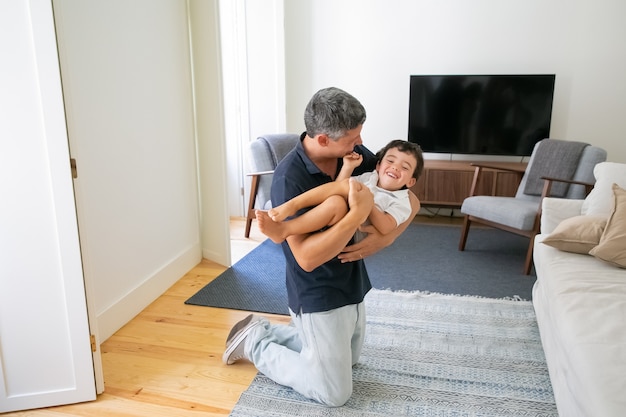 Papá feliz sosteniendo a su hijo en las manos y de rodillas en la sala de estar.