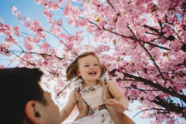 Papá feliz tiene hijita en sus brazos de pie bajo el árbol con flores