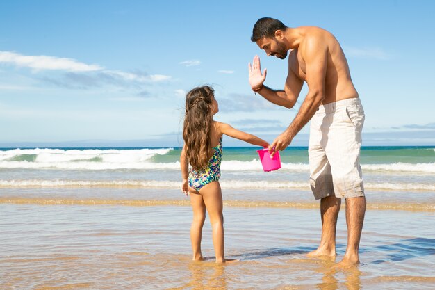 Papá guapo e hija recogiendo conchas con balde en la playa juntos, dando cinco