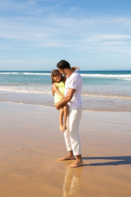 Papá joven hermoso que pasa el tiempo libre con la pequeña hija en la playa en el mar, sosteniendo al niño en brazos