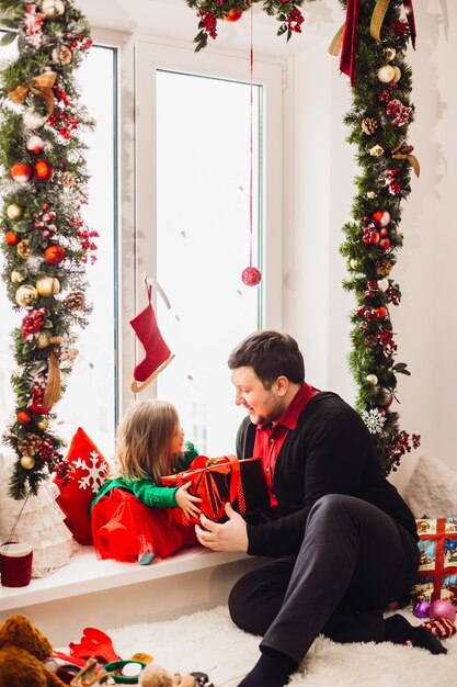 Papá juega con su hija pequeña ante una ventana brillante decorada para Navidad