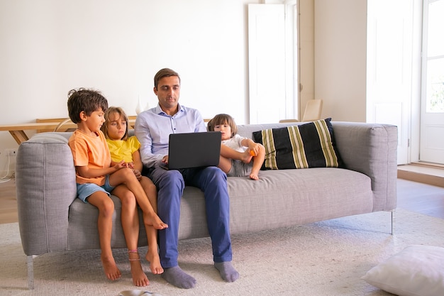 Papá de mediana edad enfocado sentado en el sofá con niños y escribiendo en la computadora portátil. Padre caucásico relajante con niños lindos en la sala de estar y viendo películas. Concepto de paternidad y tecnología digital