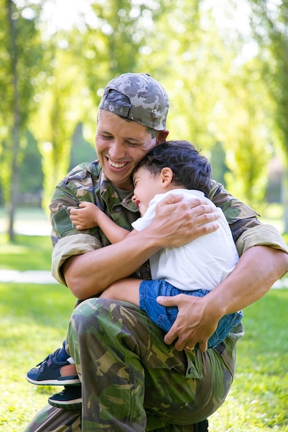 Papá militar alegre abrazando al pequeño hijo, sosteniendo al niño en brazos al aire libre después de regresar de un viaje misionero. Disparo vertical. Reunión familiar o concepto de regreso a casa