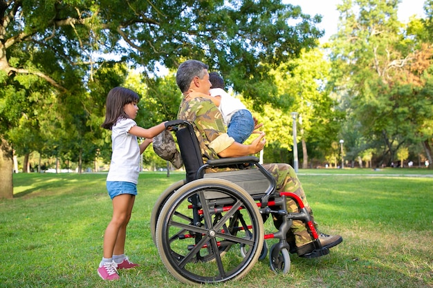 Papá militar discapacitado de mediana edad caminando con dos niños en el parque. Niña sosteniendo asas de silla de ruedas, niño de pie en el regazo de papá. Veterano de guerra o concepto de discapacidad