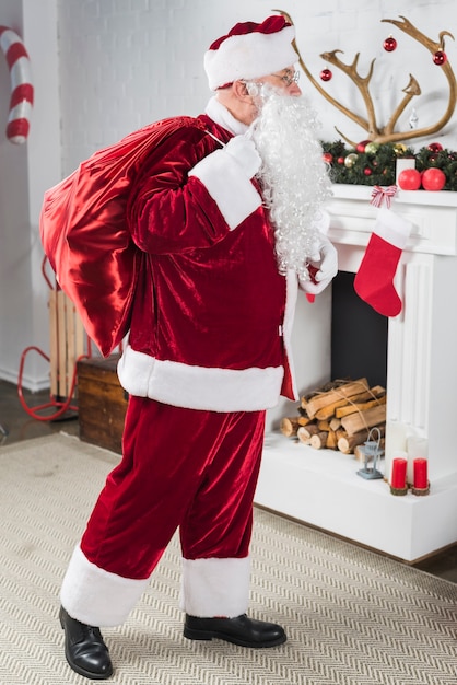 Foto gratuita papá noel con gran saco de regalos detrás de la espalda junto a la chimenea