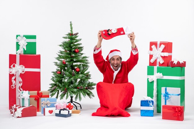 Papá Noel positivo sentado en el suelo y levantando el calcetín de Navidad en la cabeza cerca de regalos y árbol de año nuevo decorado sobre fondo blanco.