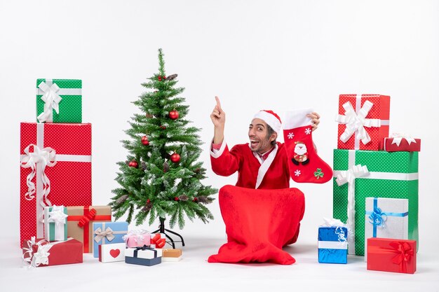 Papá Noel positivo sorprendido apuntando arriba sentado en el suelo y sosteniendo un calcetín de Navidad cerca de regalos y árbol de año nuevo decorado sobre fondo blanco