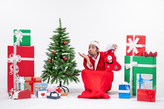 Papá Noel positivo sorprendido sentado en el suelo y sosteniendo un calcetín de Navidad cerca de regalos y árbol de año nuevo decorado sobre fondo blanco.