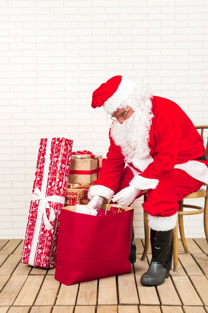 Papá Noel sentado y poniendo regalos en una bolsa de papel.
