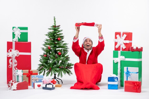 Papá Noel sonriente sentado en el suelo y levantando el calcetín de Navidad en la cabeza cerca de regalos y árbol de año nuevo decorado sobre fondo blanco.