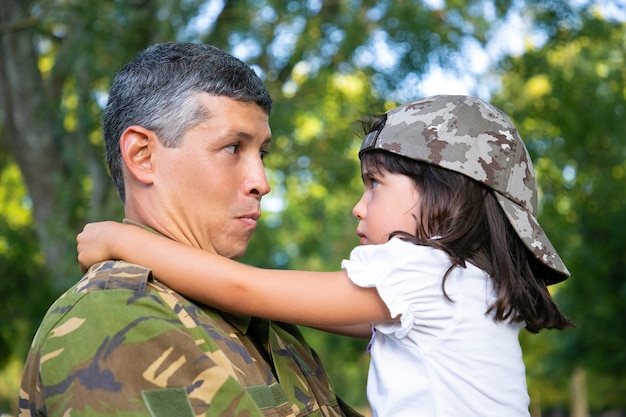 Papá positivo en uniforme de camuflaje sosteniendo a la hija ofendida en brazos, abrazando a la niña al aire libre después de regresar de un viaje misionero militar. Fotografía de cerca. Reunión familiar o concepto de regreso a casa