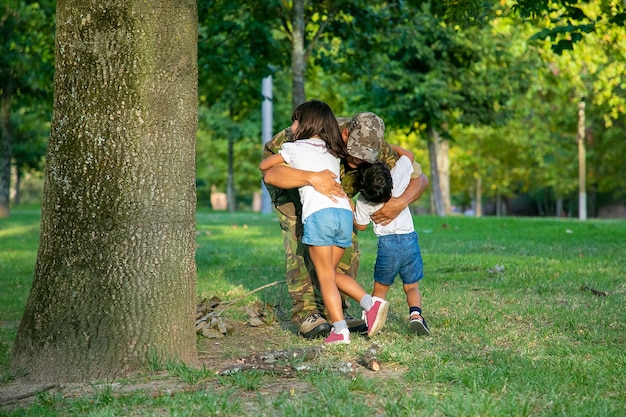 Papá reunido con dos niños después de un viaje misionero militar, abrazando a los niños en el césped del parque.