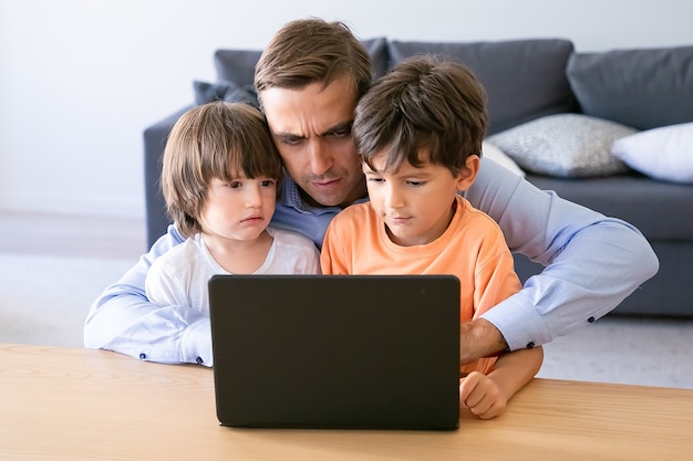 Papá serio trabajando en una computadora portátil y abrazando a sus hijos. Padre caucásico concentrado usando la computadora portátil en casa. Dos chicos guapos sentados de rodillas. Concepto de paternidad, infancia y tecnología digital.