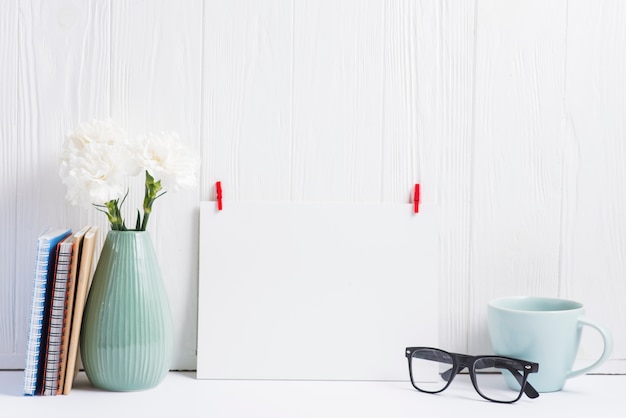 Papel blanco en blanco con clavija de ropa roja; los anteojos; vaso; Florero y libros sobre fondo de textura de madera.