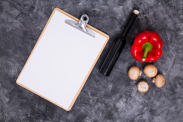 Papel blanco en blanco sobre portapapeles con pimiento; Seta y botella sobre fondo negro con textura