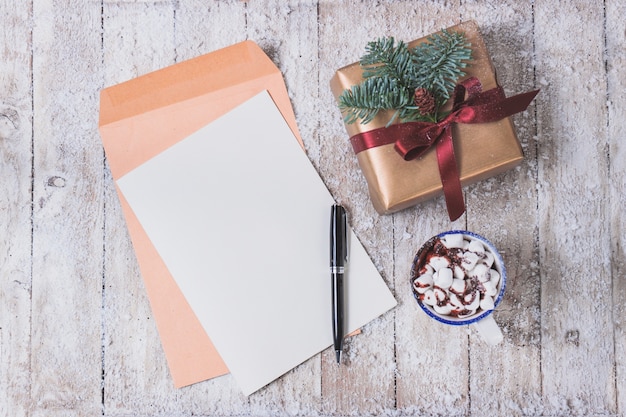 Papel en blanco con un bolígrafo y una taza con malvaviscos y un regalo