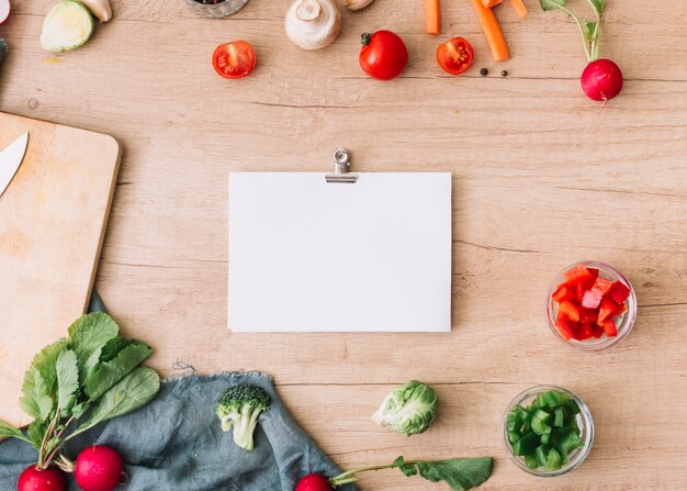 Papel en blanco con clip de papel rodeado de verduras en mesa de madera