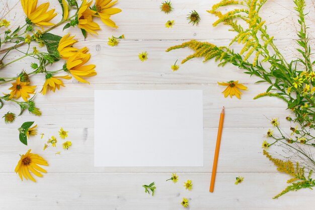 Un papel en blanco junto a un lápiz amarillo sobre una superficie de madera con flores de pétalos amarillos