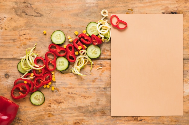 Foto gratuita papel marrón en blanco además de rebanada de pimentón; pepino y maíz en mesa de madera