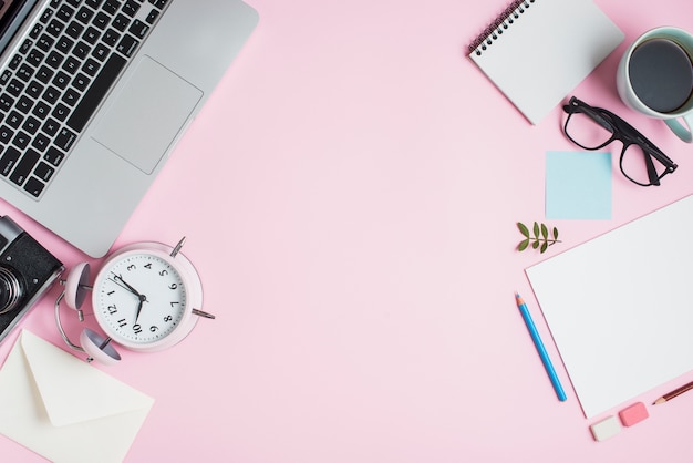 Papelerías; ordenador portátil; cámara; sobre; Reloj despertador y papelería sobre fondo rosa.