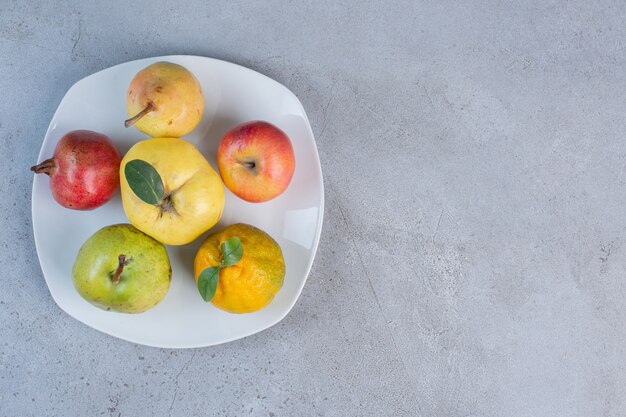 Paquete surtido de granada, peras, mandarina, membrillo y manzana en un plato sobre fondo de mármol.