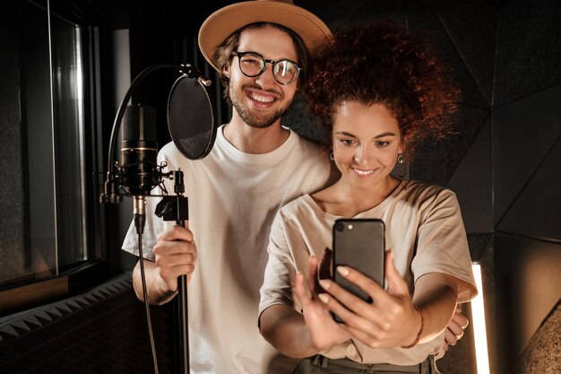 Un par de cantantes atractivos y elegantes felizmente tomándose selfie juntos en un teléfono inteligente en un moderno estudio de grabación de sonido