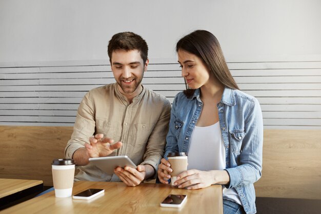 Par de entusiastas especialistas en marketing sentados a la mesa en la cafetería, sonriendo, tomando café, hablando sobre el trabajo, usando tabletas digitales y teléfonos inteligentes.