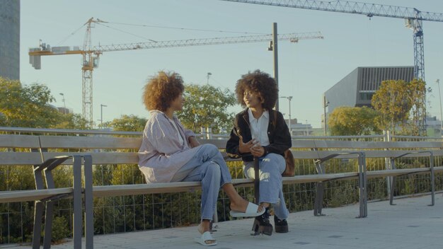 Un par de mujeres negras de raza mixta en el parque sentadas y hablando con edificios de la ciudad en el fondo