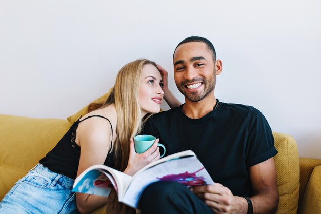 Par relajarse en el sofá en la habitación. Mujer mirando al hombre con ojos amorosos y sosteniendo una taza en la mano. El hombre está sonriendo y mirando la revista. Pasen tiempo juntos, felicidad, amor, comodidad del hogar.