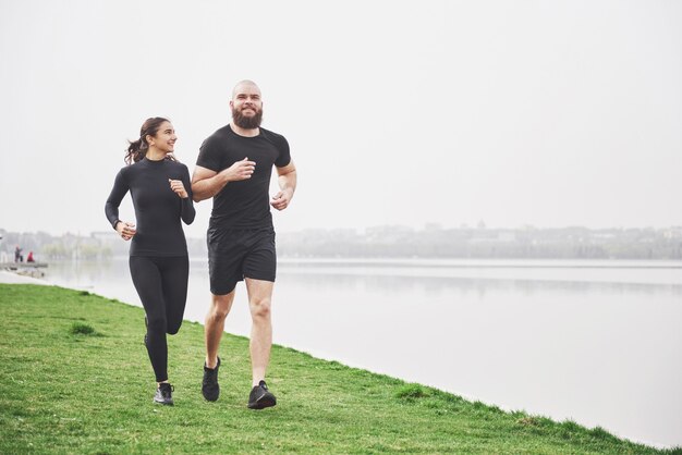 Par trotar y correr al aire libre en el parque cerca del agua. Joven barbudo hombre y mujer haciendo ejercicio juntos en la mañana