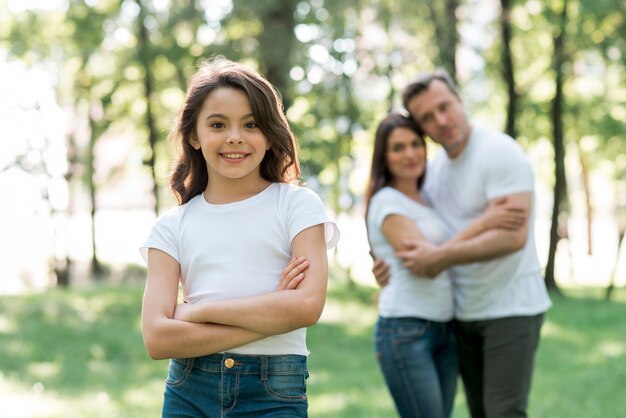 Pareja abrazándose detrás de su bonita hija mirando a cámara en el parque