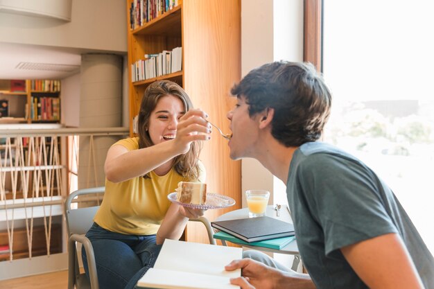 Pareja adolescente alegre comiendo pastel en la biblioteca