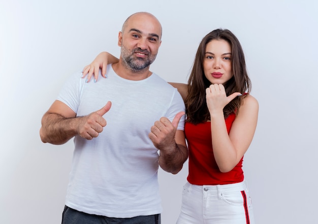 Pareja adulta hombre sonriente mostrando los pulgares hacia arriba y mujer confiada poniendo la mano sobre su hombro y apuntando al lado