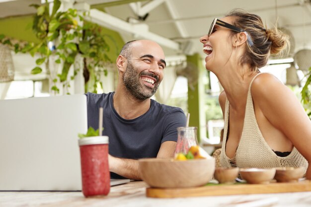 Pareja adulta relajándose en el café en la acera, bebiendo batidos, conversando animadamente y usando la computadora portátil.