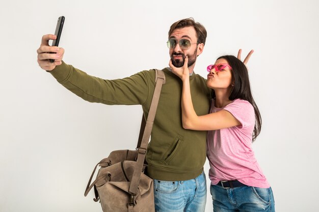 Pareja aislada, mujer muy sonriente en camiseta rosa y hombre en sudadera con bolsa de viaje, jeans y gafas de sol, divirtiéndose, viajando juntos haciendo una foto divertida selfie en el teléfono