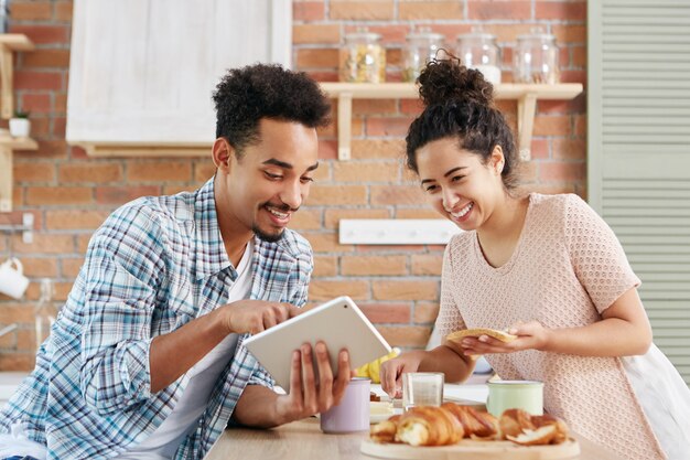 Pareja alegre elige muebles nuevos en la cocina, mira felizmente en la pantalla de la tableta