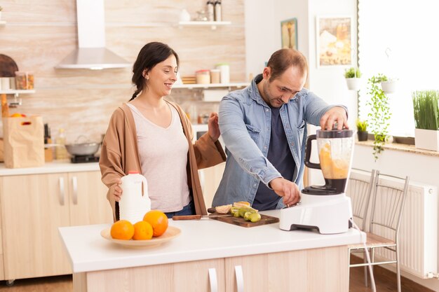 Pareja alegre prepara batido con licuadora. Esposa sosteniendo la botella de leche en la cocina. Estilo de vida saludable, despreocupado y alegre, comiendo dieta y preparando el desayuno en una acogedora mañana soleada