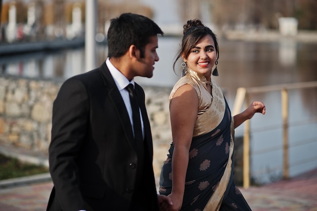 Pareja de amigos indios elegantes y de moda de mujer en sari y hombre en traje caminando juntos y tomados de la mano
