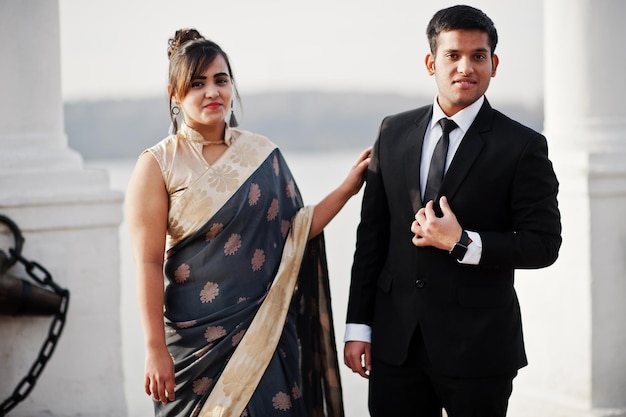 Pareja de amigos indios elegantes y de moda de mujer en sari y hombre en traje posaron en la orilla del puerto deportivo