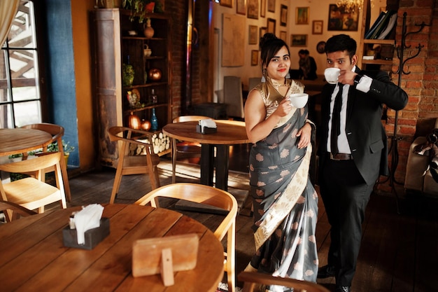 Pareja de amigos indios elegantes y de moda de mujer en sari y hombre en traje sentado en el café y bebiendo té