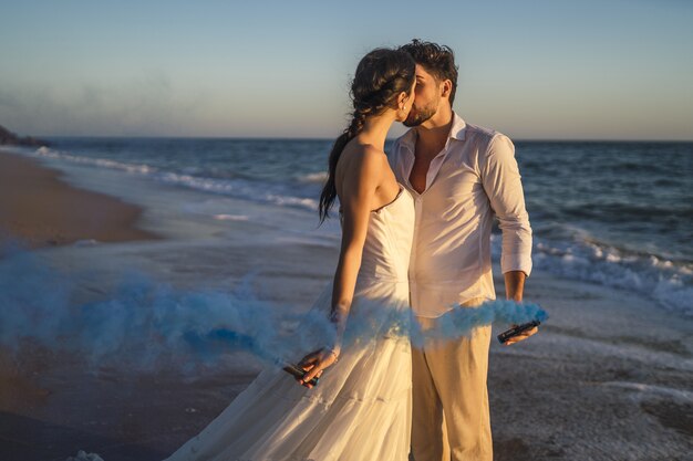 Pareja amorosa caucásica sosteniendo un humo de color azul y besándose en la playa durante una boda