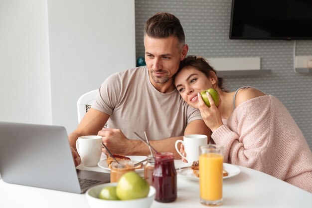 Pareja amorosa contenta desayunando mientras está sentado a la mesa y usando la computadora portátil en la cocina