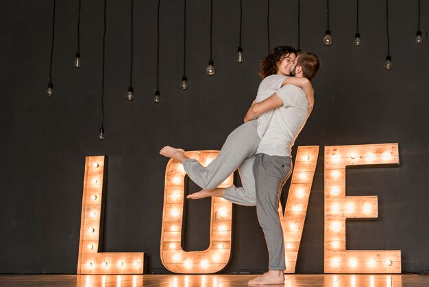 Pareja amorosa de pie delante de un texto de amor iluminado contra una pared negra