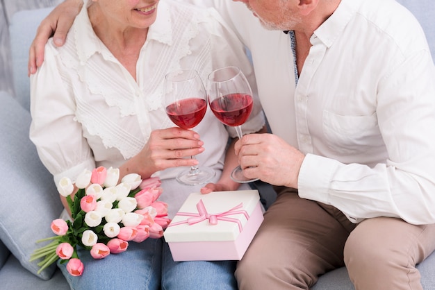 Foto gratuita pareja amorosa sentado en el sofá con copas de vino; caja de regalo y ramo de flores de tulipán.