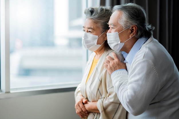 Pareja de ancianos jubilados asiáticos con máscaras faciales miran el mundo a través de la ventana de su hogar abrazo de mano y expresión de tristeza solitaria personas asiáticas aislamiento en el hogar distanciamiento social quedarse en casa concepto de ideas