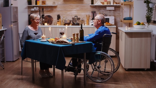 Pareja de ancianos regresando a casa para una cena romántica. Anciano en silla de ruedas cenando con su esposa alegre sentado en la mesa de la cocina. Marido discapacitado paralizado inmovilizado con cena romántica