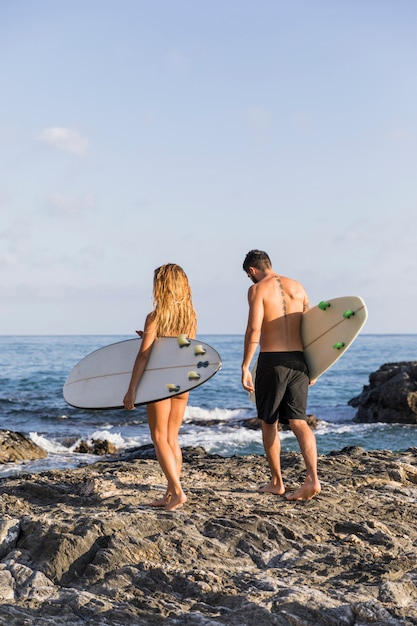 Pareja anónima con tablas de surf caminando en la costa rocosa